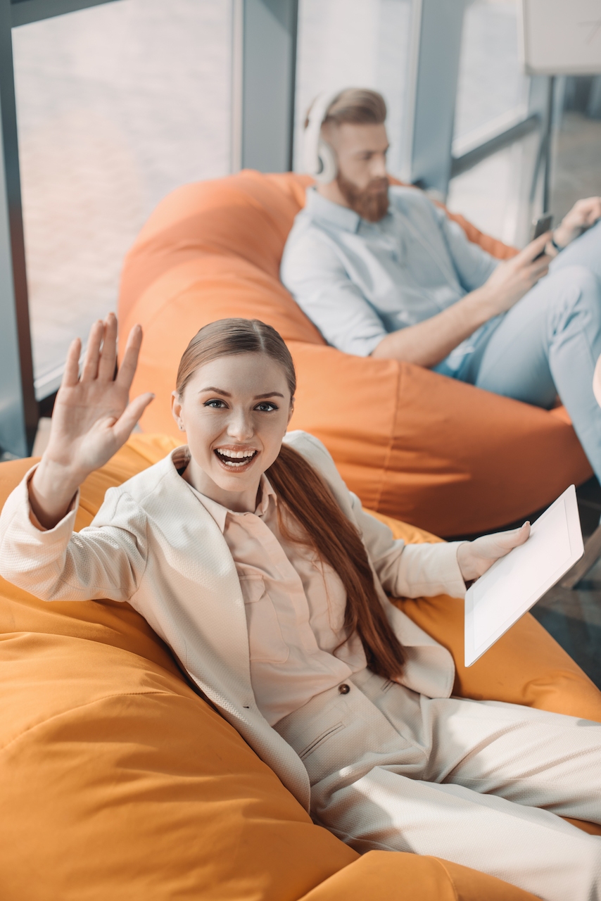 Young smiling businesswoman waving at camera with businessman sitting behind
